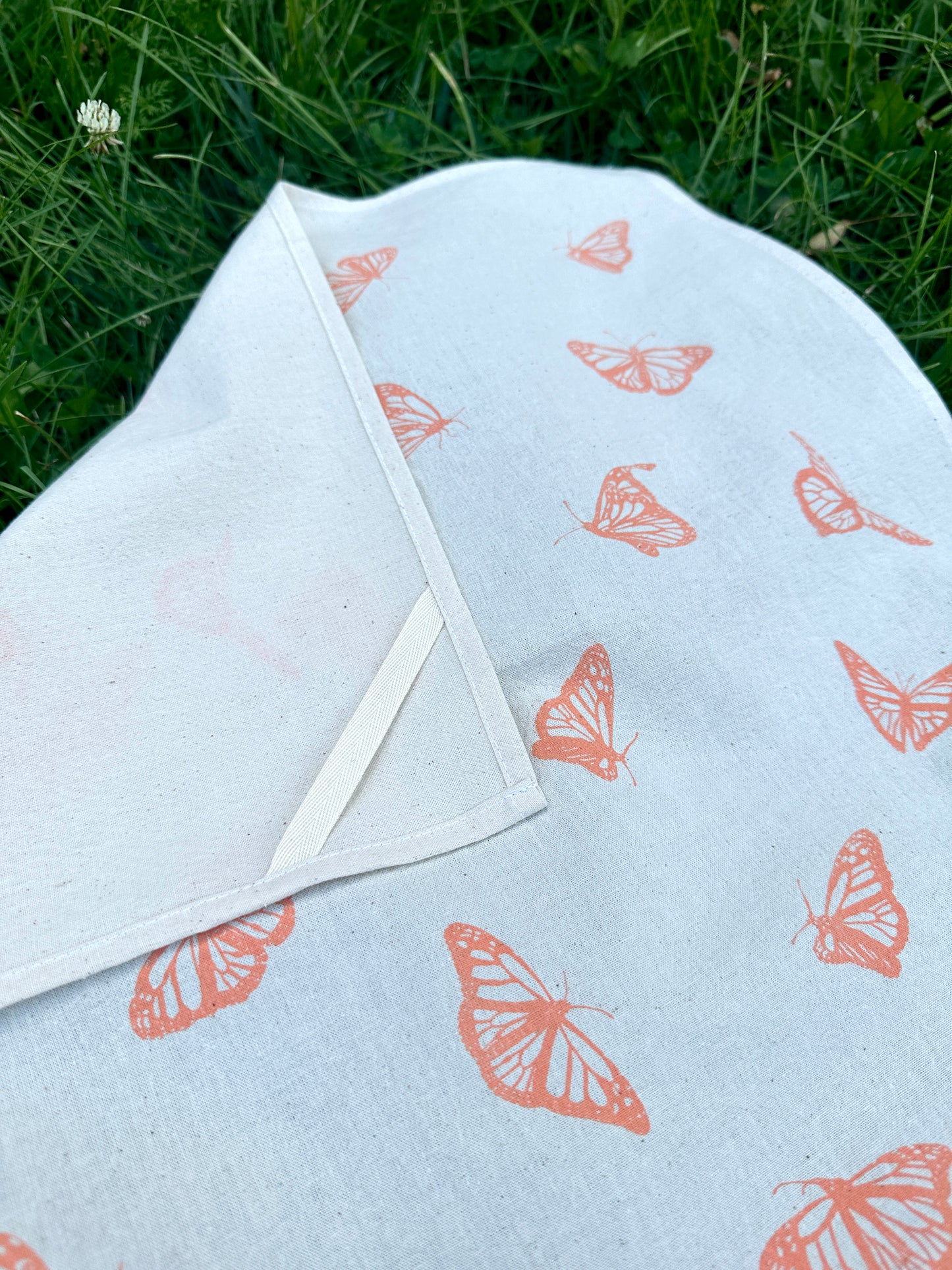 Tea towel with a monarch butterfly pattern, laid flat on grass, with a corner folded over showing the loop on the back corner of the towel.